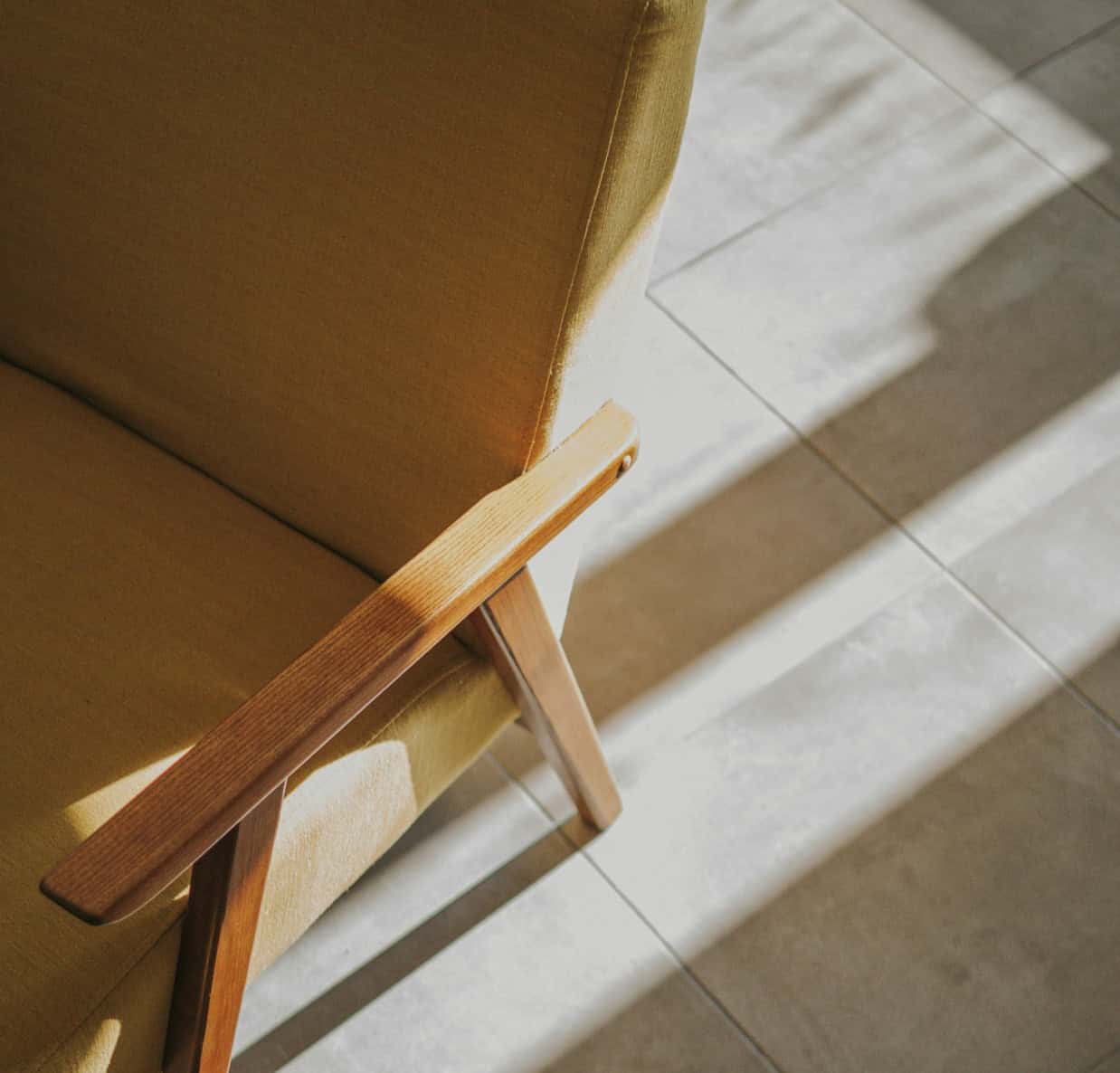 Armchair with wooden armrest and yellow upholstery, illuminated by natural sunlight on tiled floor.